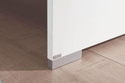 Nahaufnahme einer weißen Möbelkante mit grauem Sockel auf Holzboden, perspektivisch von der Seite aufgenommen.