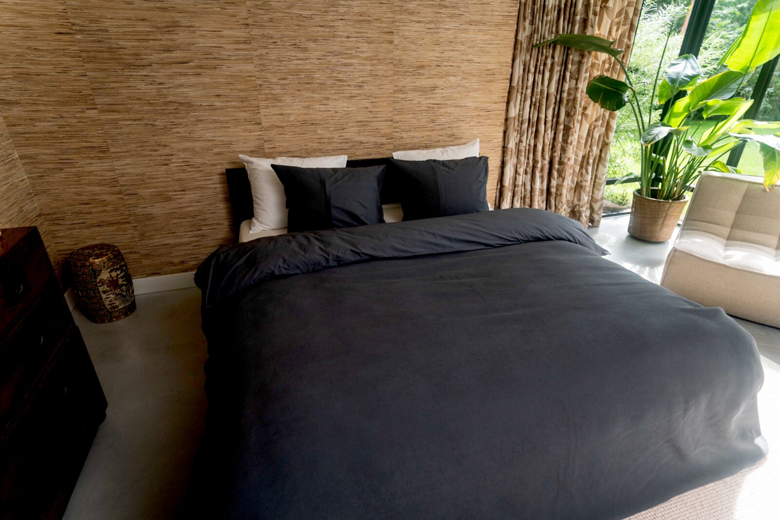Blick auf ein stilvoll eingerichtetes Schlafzimmer mit einem Bett, das mit dunkelgrauer Bettwäsche in der Größe 135x200cm bezogen ist. Im Hintergrund sind eine beige Wand, Vorhänge und eine große grüne Pflanze zu sehen.