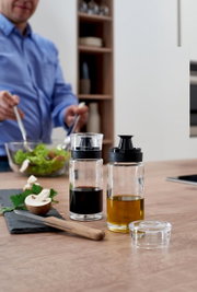 Essig- und Ölflaschen Cucina auf einem Küchentisch, im Hintergrund eine Person beim Zubereiten eines Salats, seitliche Perspektive.
