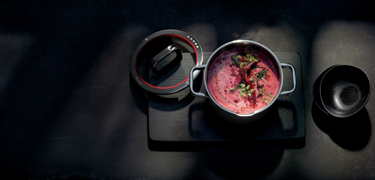 Top-Down-Ansicht eines Gemüsetopfs Function 4 mit roter Suppe und Kräutern, daneben ein abgenommener Deckel und eine schwarze Schale.
