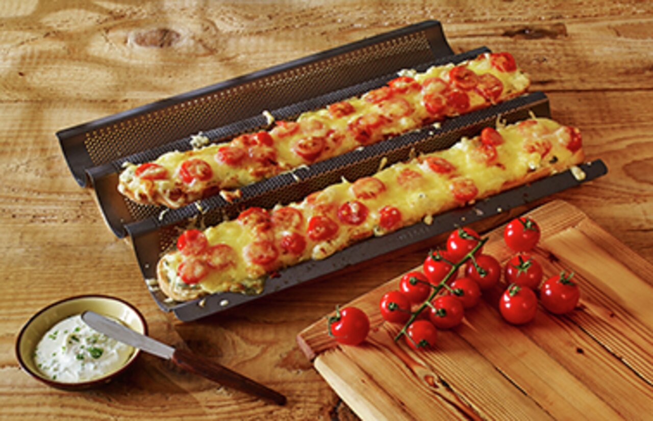 Een bakplaat met twee belegde baguettes, van bovenaf gefotografeerd, op een houten tafel met tomaten en dip.
