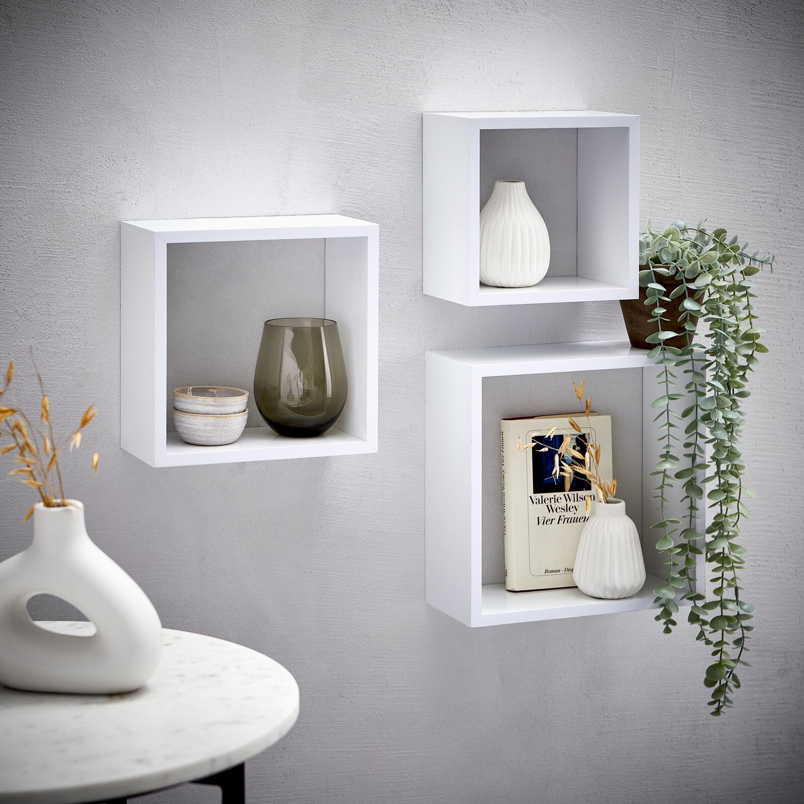 XORA Wandregal-3er-Set TRIO Dreiteiliges Wandregal-Set in Weiß, dekoriert mit Vasen, einem Buch und einer hängenden Pflanze, aus seitlicher Perspektive fotografiert.