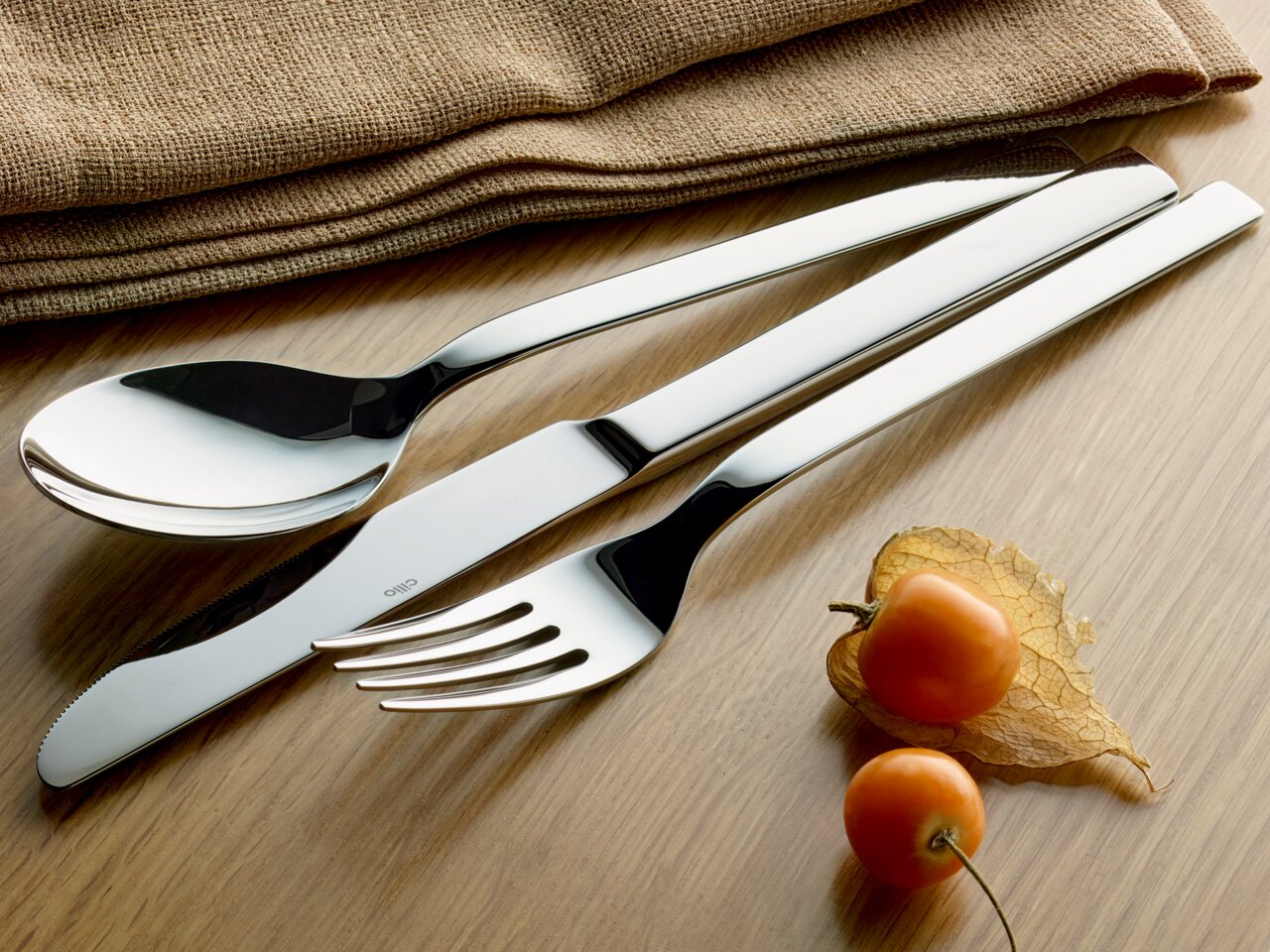Elegantes Besteck-Set bestehend aus Löffel, Messer und Gabel auf einem Holztisch, mit dekorativen Physalis und Stoffservietten im Hintergrund, aus der Vogelperspektive fotografiert.
