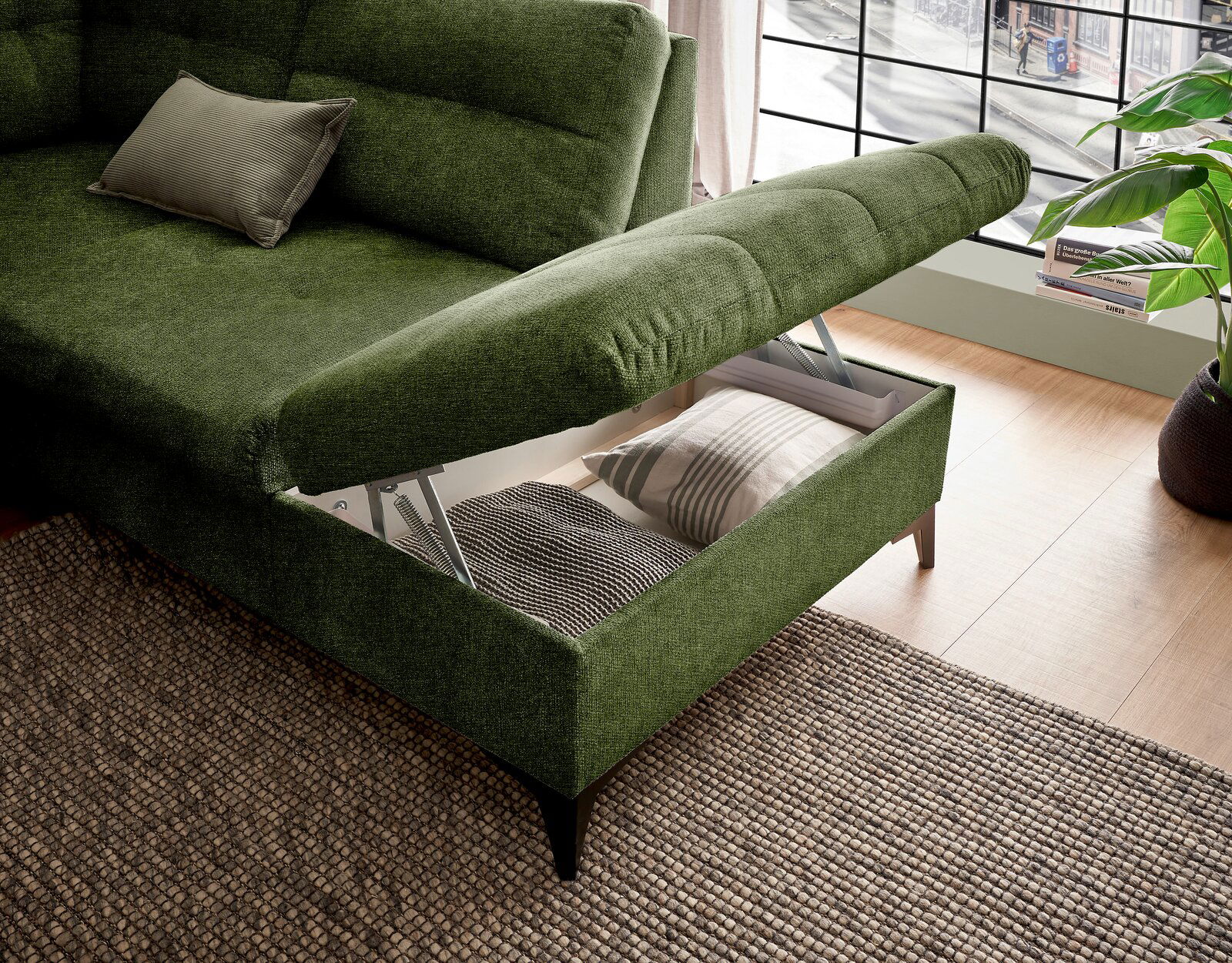 Grünes Ecksofa mit Stauraum, geöffnet, von schräg oben fotografiert, zeigt Kissen und Decke im Stauraum.
