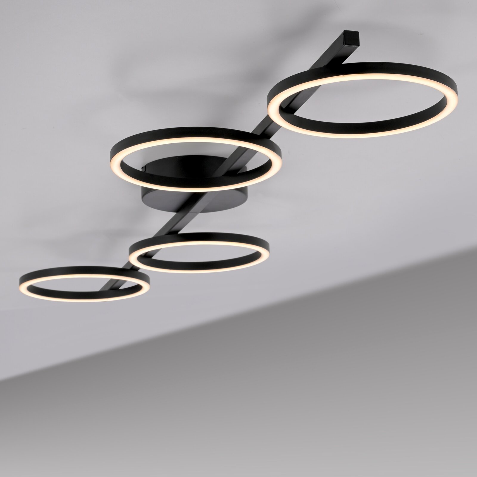 Moderne schwarze LED-Deckenleuchte mit vier ringförmigen Lichtkreisen an einer geraden Schiene, warmweiß leuchtend, fotografiert aus der Untersicht in schräger Perspektive.