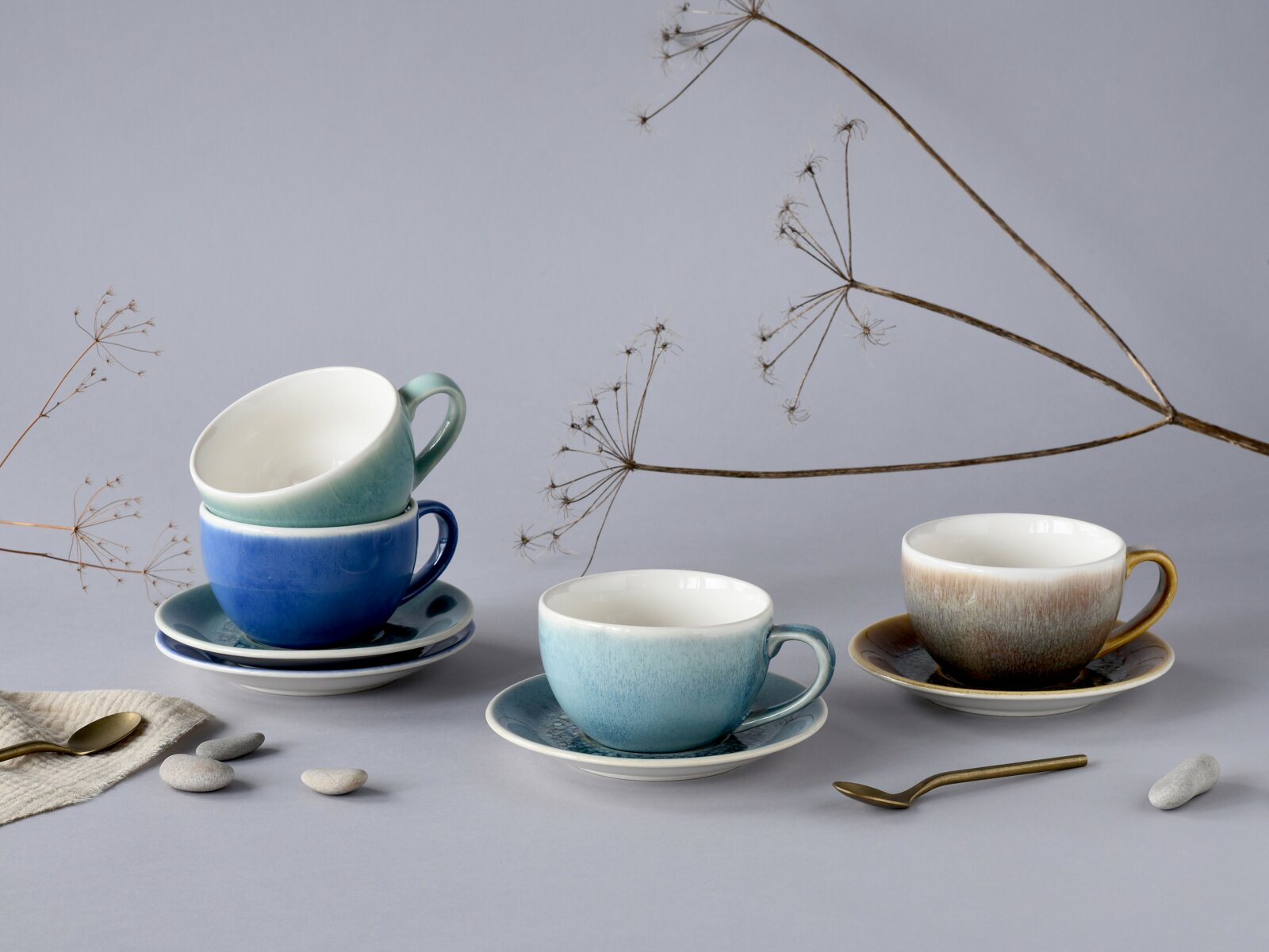 Creatable Cappuccino-Set 8-tlg CALDERA MIX Stillleben aus Keramik-Kaffeetassen mit Untertassen in Blau-, Türkis- und Brauntönen auf grauem Hintergrund, teils gestapelt, mit Löffeln, Steinen und Trockenblumen; Perspektive: leicht erhöhte Frontansicht.