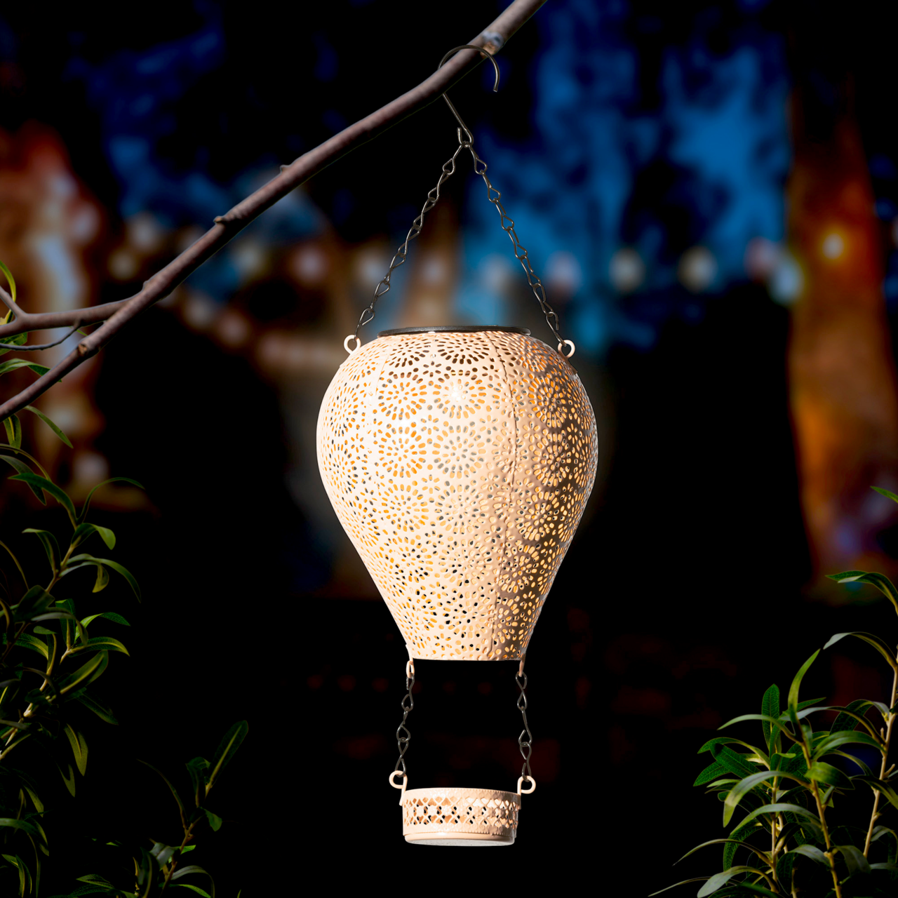 Metallischer Solarballon in Form eines Heißluftballons, aufgehängt an einem Ast, mit filigranen Mustern, die im Dunkeln leuchten. Perspektive von der Seite.