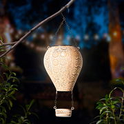 Metallischer Solarballon in Form eines Heißluftballons, aufgehängt an einem Ast, mit filigranen Mustern, die im Dunkeln leuchten. Perspektive von der Seite.