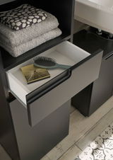 Hochschrank mit offener Schublade, gefüllt mit einer Haarbürste und einem goldenen Accessoire, daneben gestapelte Handtücher. Perspektive von oben.