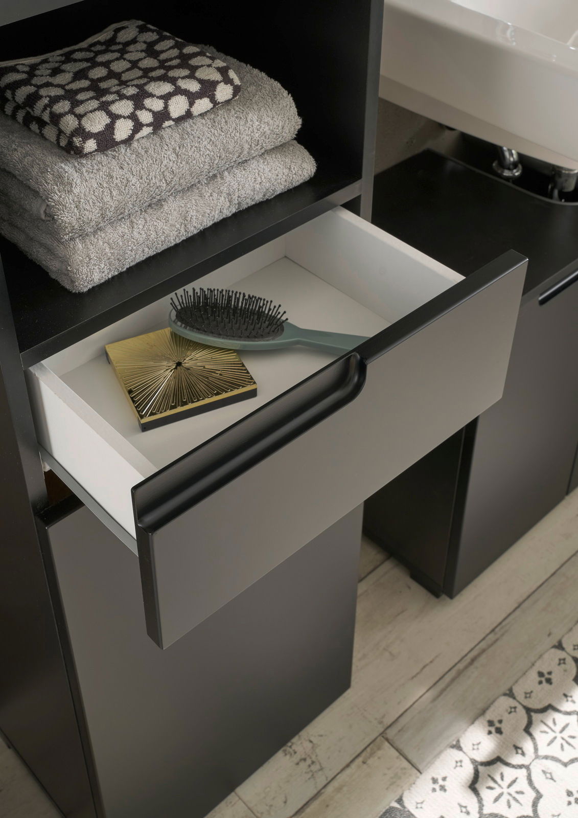 Hochschrank mit offener Schublade, gefüllt mit einer Haarbürste und einem goldenen Accessoire, daneben gestapelte Handtücher. Perspektive von oben.