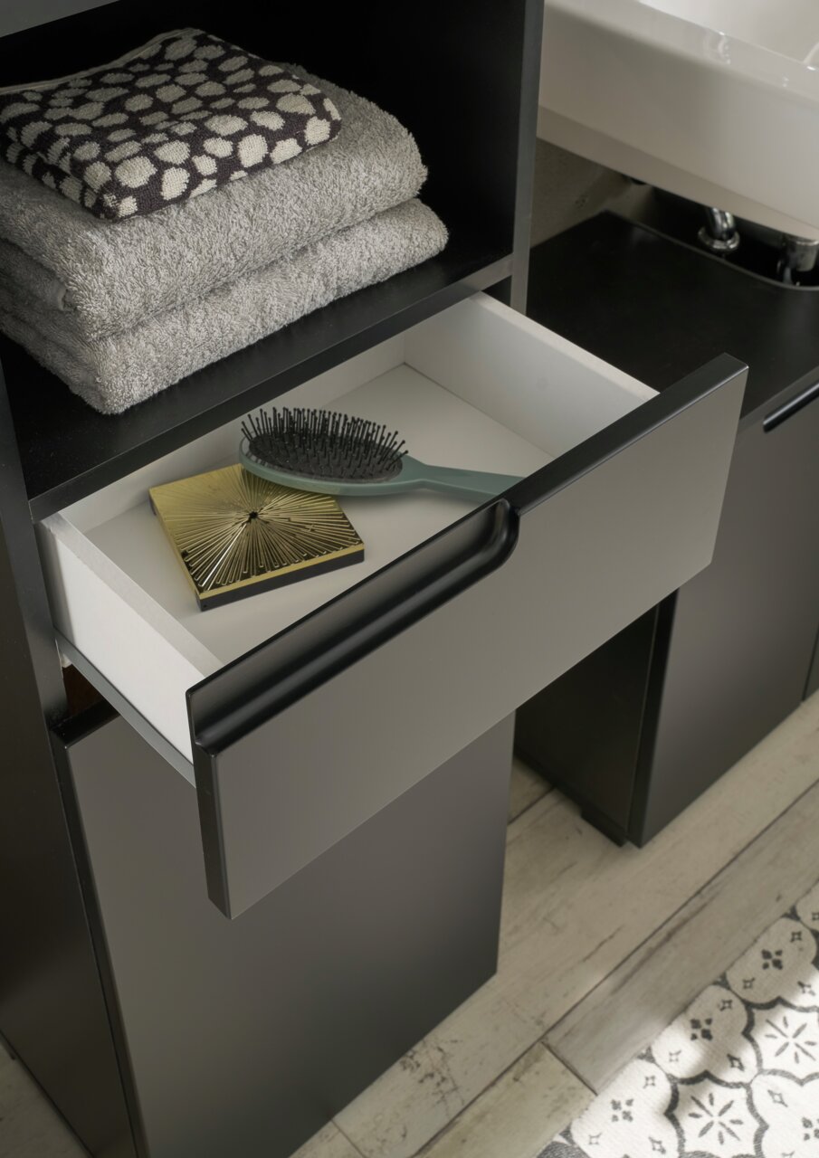 Hochschrank mit offener Schublade, gefüllt mit einer Haarbürste und einem goldenen Accessoire, daneben gestapelte Handtücher. Perspektive von oben.