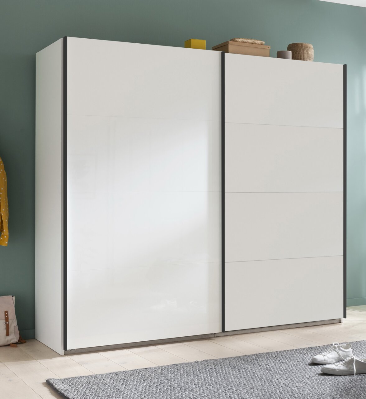Moderner weißer Schwebetürenschrank mit zwei Türen, seitliche Perspektive, auf hellem Holzboden vor einer grünen Wand.