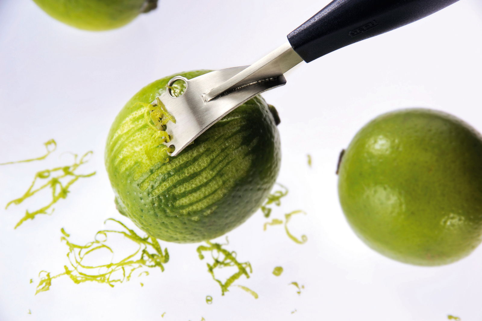 Nahaufnahme eines Zestenreißers, der die Schale einer Limette abzieht, mit herabfallenden Zesten. Perspektive von oben.