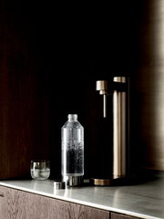 Brus Wassersprudler in dunklem Braun auf einer Küchenarbeitsplatte, mit einer gefüllten Sprudelflasche und einem Glas, seitliche Perspektive.