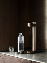 Brus Wassersprudler in dunklem Braun auf einer Küchenarbeitsplatte, mit einer gefüllten Sprudelflasche und einem Glas, seitliche Perspektive.