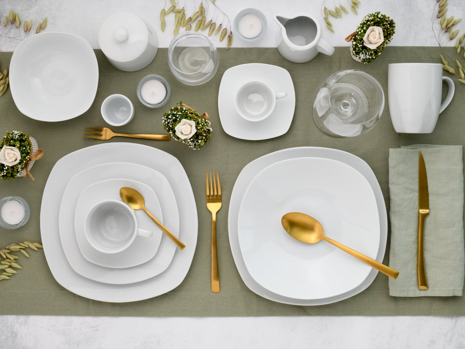 Draufsicht auf modern gedeckten Tisch mit weißem quadratischem Porzellan-Geschirr (Teller, Schalen, Tassen), Glaswaren und goldfarbenem Besteck auf grünem Tischläufer, dekoriert mit Kerzen und kleinen Blumenarrangements