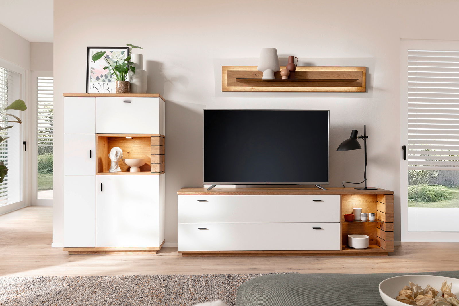 Novel Wandkombination MANITOBA Moderne Wandkombination in einem hellen Wohnzimmer, bestehend aus einem weißen Schrank mit Holzdetails und einem TV-Schrank mit integriertem Regal. Perspektive von vorne.