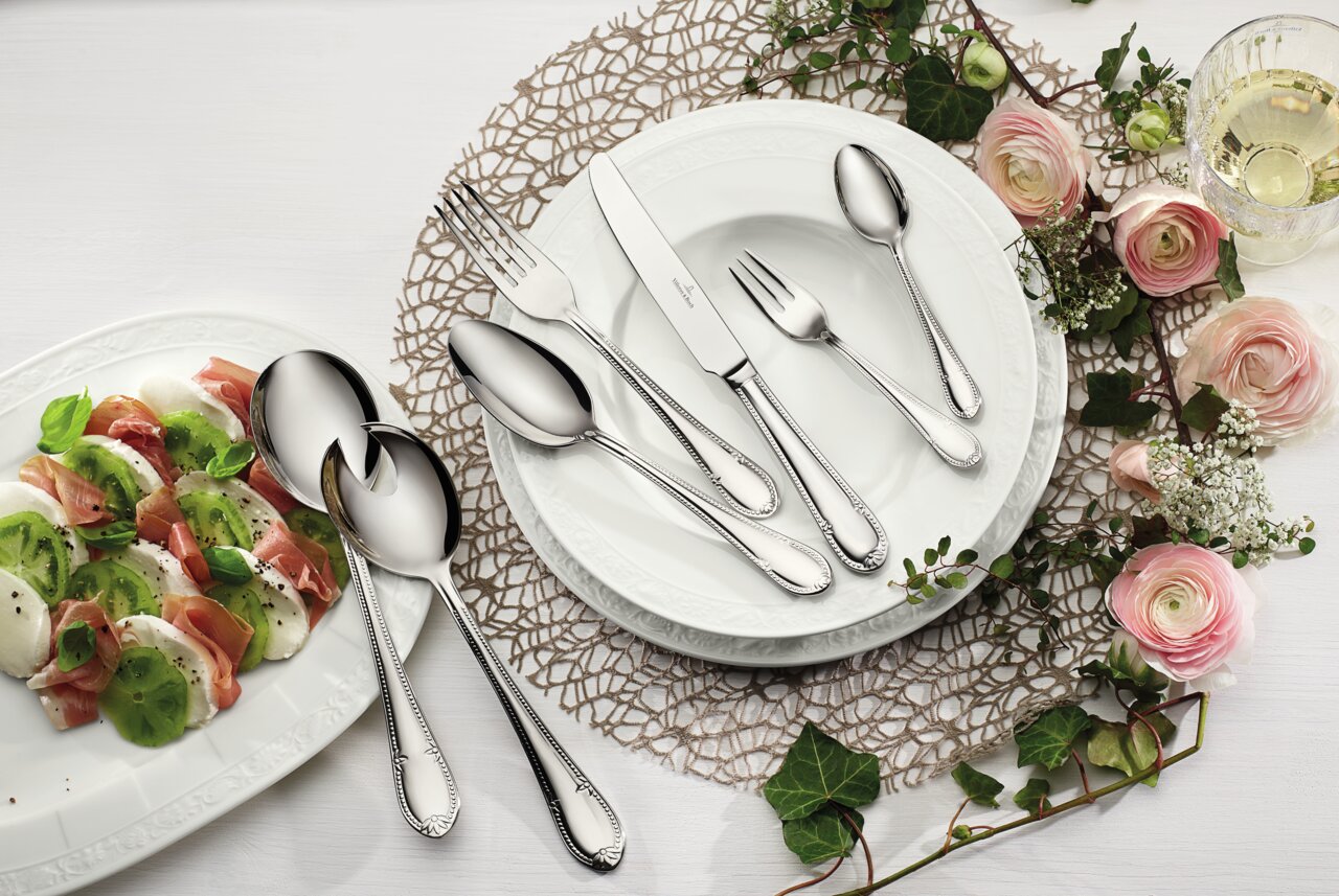 Villeroy & Boch Tafelbesteck 68-tlg. MADEMOISELLE Elegantes 68-teiliges Tafelbesteck-Set aus Edelstahl, präsentiert auf einem gedeckten Tisch mit weißem Porzellan, Blumen und einem Glas Weißwein, aus der Vogelperspektive.