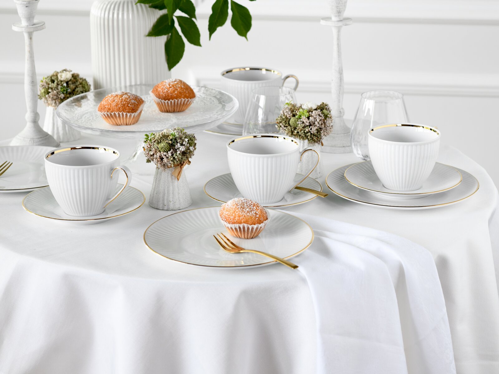 Schick gedeckter Tisch mit weißem geripptem Porzellan-Kaffeeservice mit Goldrand (Tassen, Untertassen und Dessertteller), goldfarbenem Besteck und Muffins; Perspektive: leicht erhöhte Frontansicht schräg von vorn.