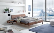 Modernes Bettgestell aus Holz in einem hellen Schlafzimmer mit Blick auf eine verschneite Berglandschaft durch große Fenster, seitliche Perspektive.
