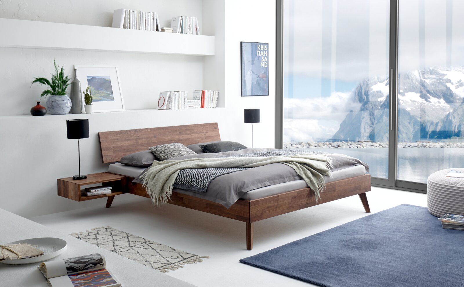Modernes Bettgestell aus Holz in einem hellen Schlafzimmer mit Blick auf eine verschneite Berglandschaft durch große Fenster, seitliche Perspektive.
