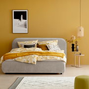 Graues Polsterbett LISSABON in einem modern eingerichteten Schlafzimmer mit gelber Wand, dekoriert mit gemusterten Kissen und Decken, seitliche Perspektive.