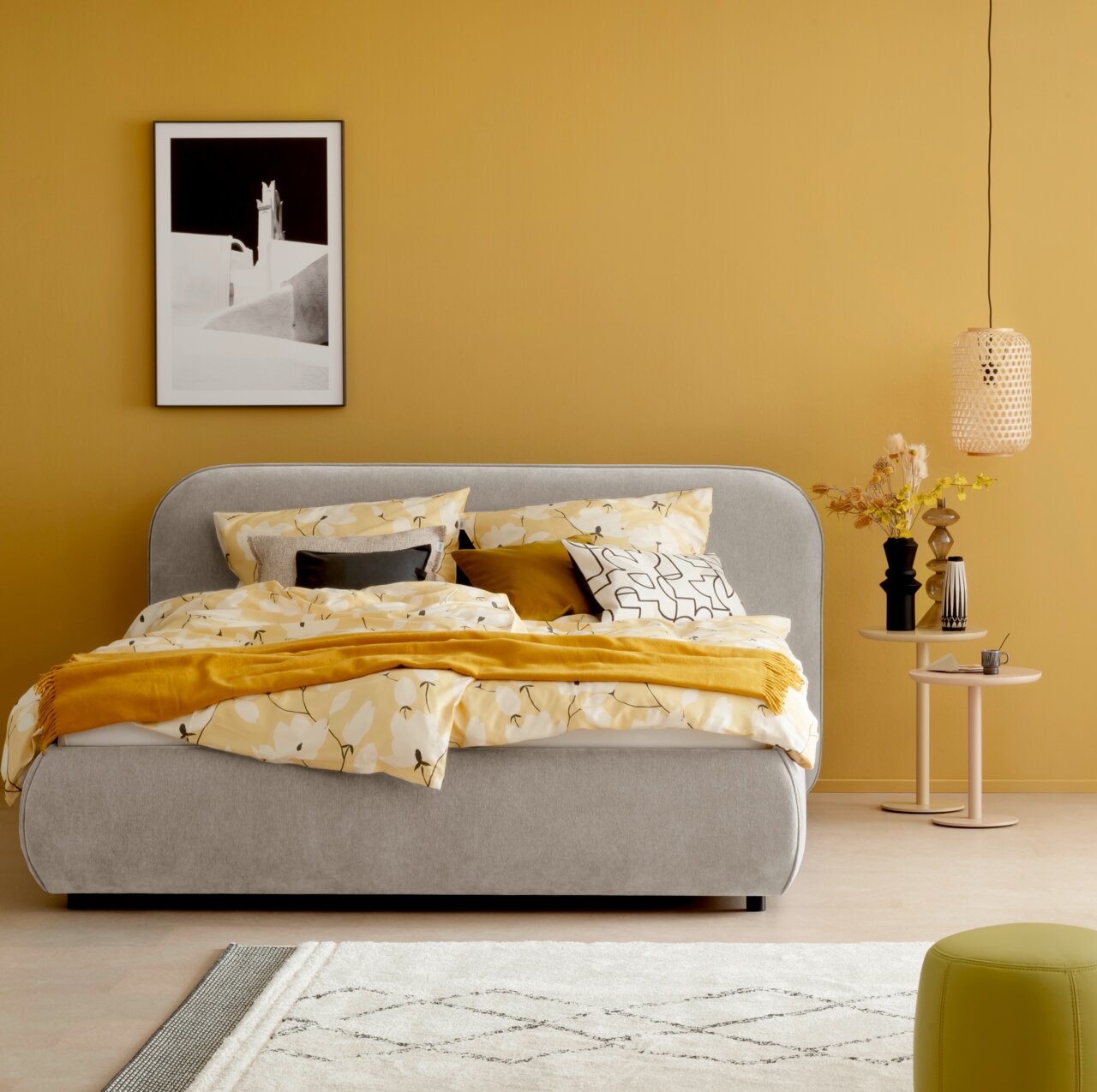 Graues Polsterbett LISSABON in einem modern eingerichteten Schlafzimmer mit gelber Wand, dekoriert mit gemusterten Kissen und Decken, seitliche Perspektive.