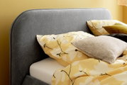 Graues Polsterbettgestell mit gelber Blumenbettwäsche und Kissen, seitliche Perspektive