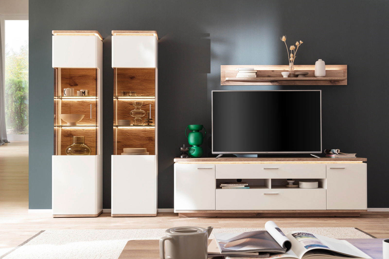 Moderne Wandkombination mit zwei beleuchteten Vitrinen, einem TV-Schrank und einem Wandregal in einem stilvollen Wohnzimmer, frontal betrachtet.
