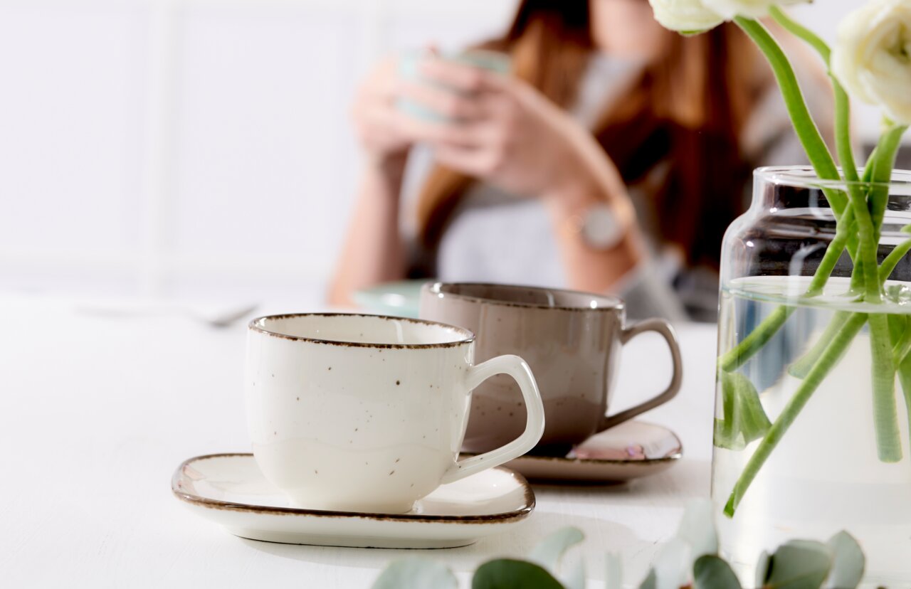 Espressountertasse Casa beige mit Tasse, seitliche Perspektive, im Hintergrund unscharfe Person und Blumen in Vase