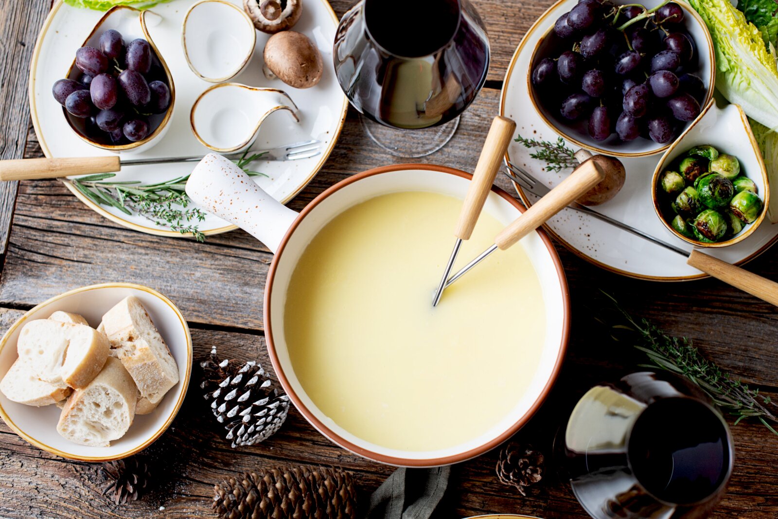 Draufsicht auf ein rustikales Käsefondue-Set: großer Topf mit geschmolzenem Käse und Fonduegabeln, dazu Brotstücke, Trauben, Rosenkohl, Champignons, Kräuter und Gläser Rotwein auf Holztisch