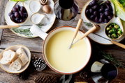 Draufsicht auf ein rustikales Käsefondue-Set: großer Topf mit geschmolzenem Käse und Fonduegabeln, dazu Brotstücke, Trauben, Rosenkohl, Champignons, Kräuter und Gläser Rotwein auf Holztisch
