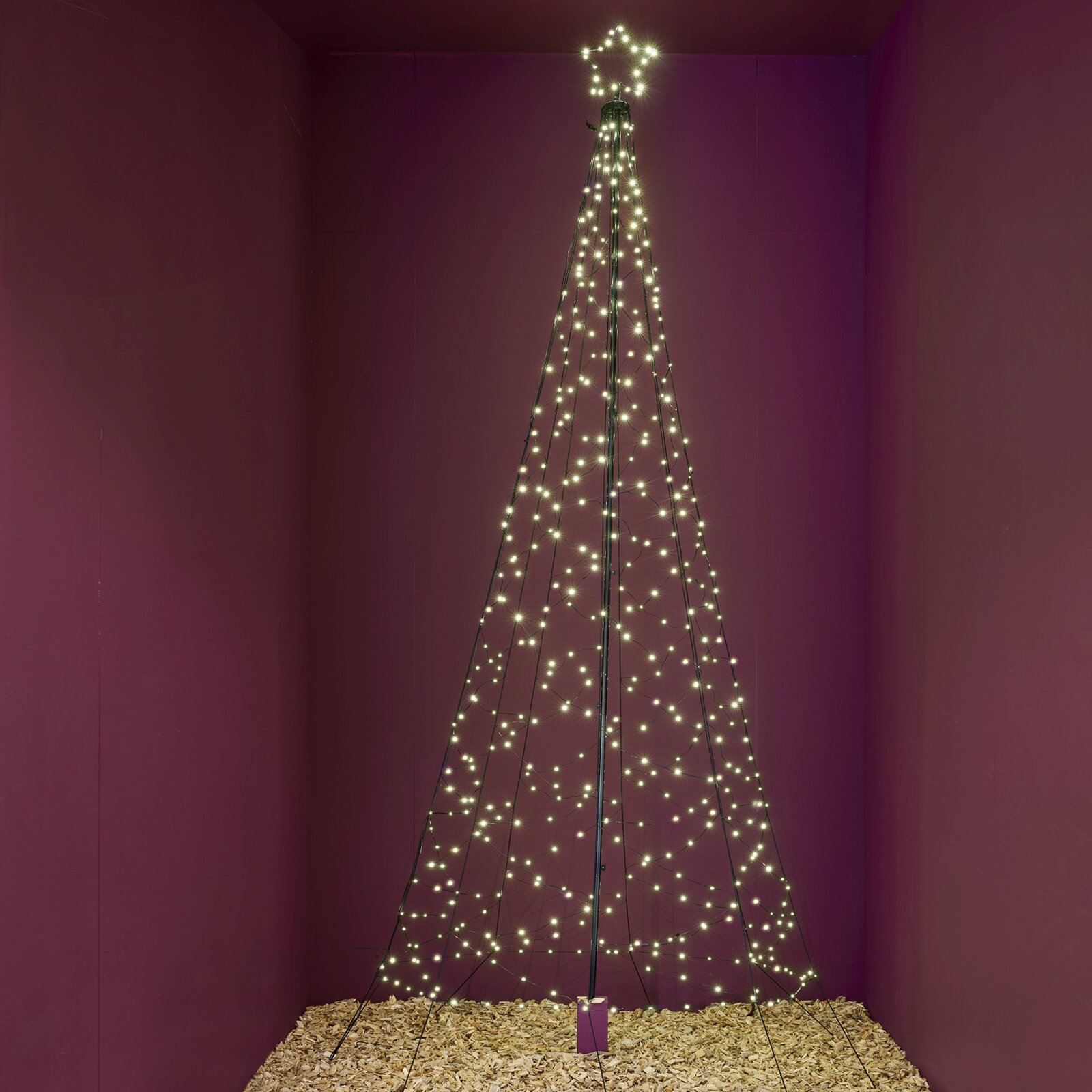 Frontaal aanzicht van een hoge, kegelvormige metalen kerstboom met warmwitte lichtjes en een verlichte ster tegen een bordeauxrode achtergrond
