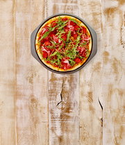 Pizzablech mit einer frisch gebackenen Pizza, belegt mit Rucola, Tomaten und Schinken, aus der Vogelperspektive auf einem rustikalen Holztisch.
