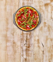 Pizzablech mit einer frisch gebackenen Pizza, belegt mit Rucola, Tomaten und Schinken, aus der Vogelperspektive auf einem rustikalen Holztisch.