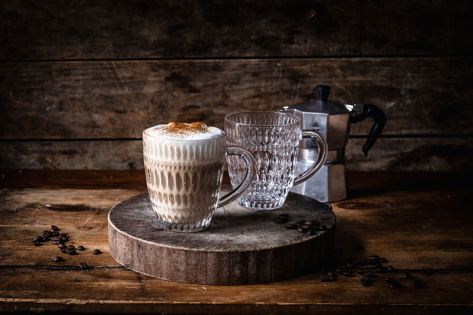 Zwei Ethno-Becher auf einem rustikalen Holzbrett, einer gefüllt mit Cappuccino, im Hintergrund eine Espressokanne. Perspektive von vorne.