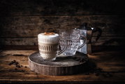 Zwei Ethno-Becher auf einem rustikalen Holzbrett, einer gefüllt mit Cappuccino, im Hintergrund eine Espressokanne. Perspektive von vorne.