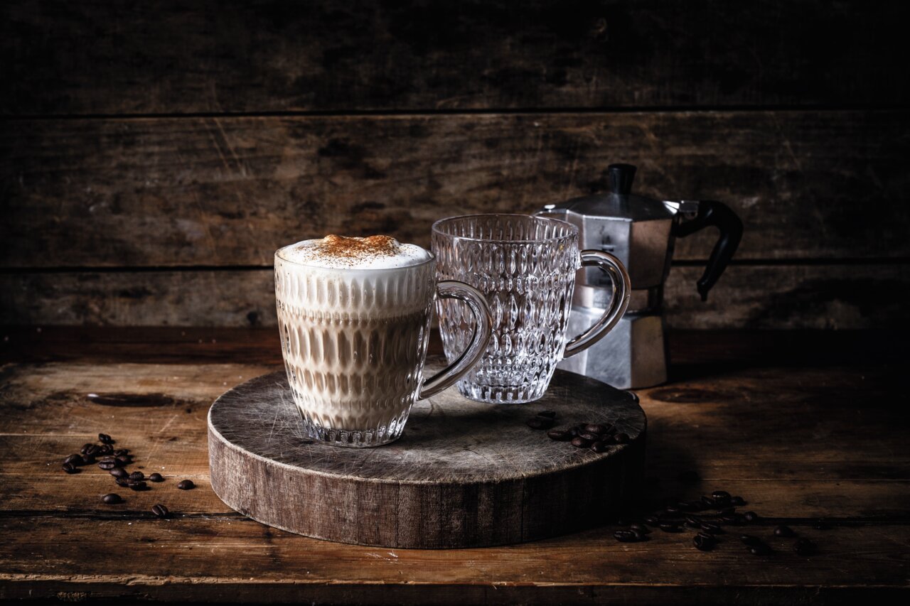 Twee etnische mokken op een rustieke houten plank, waarvan één gevuld met cappuccino, op de achtergrond een espressopot. Vooraanzicht.