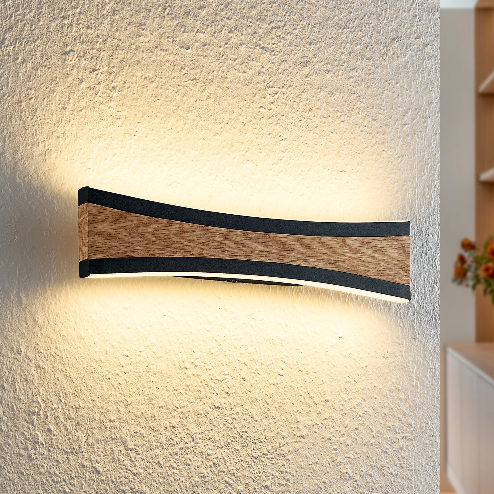 Novel LED-Wandleuchte Gebogene LED-Wandleuchte mit Holzoptik und schwarzem Rahmen an strukturierter Wand, warmweißes indirektes Licht nach oben und unten, frontale Seitenansicht.