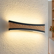 Gebogene LED-Wandleuchte mit Holzoptik und schwarzem Rahmen an strukturierter Wand, warmweißes indirektes Licht nach oben und unten, frontale Seitenansicht.
