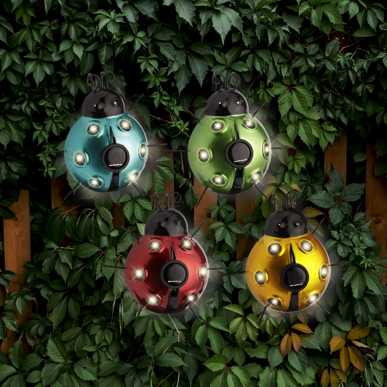 Vier bunte Solar-Insekten aus Eisen in den Farben Blau, Grün, Rot und Gelb, aufgehängt vor einem Hintergrund aus grünen Blättern, aus der Vorderansicht fotografiert.