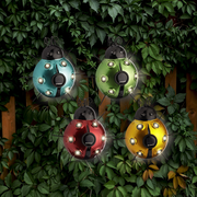 Vier bunte Solar-Insekten aus Eisen in den Farben Blau, Grün, Rot und Gelb, aufgehängt vor einem Hintergrund aus grünen Blättern, aus der Vorderansicht fotografiert.