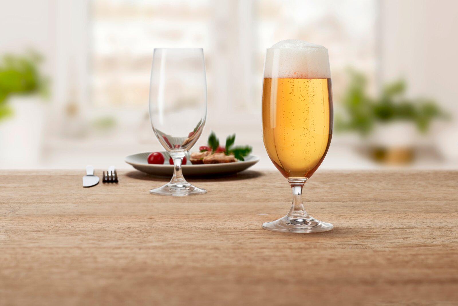 Bierglas mit Schaumkrone, gefüllt mit goldenem Bier, auf einem Holztisch im Vordergrund, mit einem leeren Glas und einem Teller mit Snacks im Hintergrund.