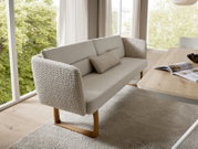 Beige gestoffeerde eetkamerbank met hoge rugleuning, decoratief kussen, gestructureerde armleuningen en houten sledepoten, schuin van voren gezien in een lichte woonruimte