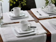 Weißes Porzellan-Kaffeegeschirr mit Tasse auf quadratischer Untertasse und Teller, auf gedecktem Holztisch mit weißer Serviette; Perspektive: leicht erhöhte, schräge Ansicht von vorne