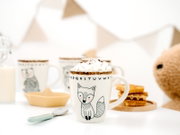 Weiße Keramiktasse mit niedlichem Fuchsmotiv und Buchstabenrand, gefüllt mit Kakao und Mini-Marshmallows, frontal aus leichter Vogelperspektive in gemütlicher Frühstücksszene