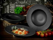 Schwarzes, geriffeltes Teller- und Schüssel-Set aus Keramik/Porzellan mit Pastaschale und Speisetellern auf rustikalem Holztisch, fotografiert in schräger Vorderansicht.