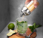 Hand hält einen Zucker- oder Milchdosierer über einem Glas mit Minzblättern, daneben Limettenscheiben und ein Löffel auf einem Holzbrett. Perspektive von schräg oben.