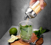 Hand hält einen Zucker- oder Milchdosierer über einem Glas mit Minzblättern, daneben Limettenscheiben und ein Löffel auf einem Holzbrett. Perspektive von schräg oben.