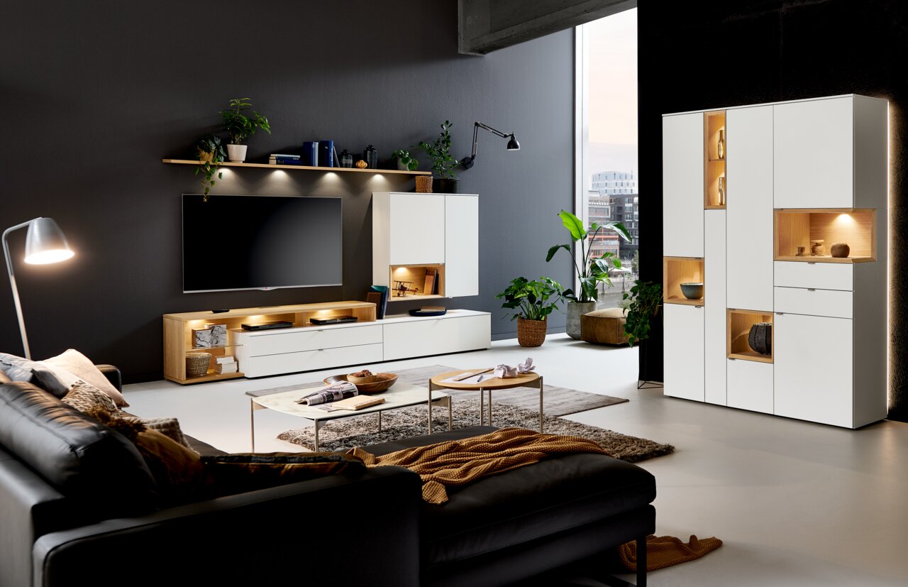 Moderne Wohnzimmermöbel mit weißem Highboard, Sideboard und Wandboard, LED-Beleuchtung, Pflanzen und Dekorationen, seitliche Perspektive.